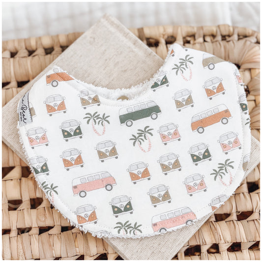 Unisex baby bib with a print of retro vans and palm trees in muted pastel tones on a soft white background, laid flat on a woven basket.