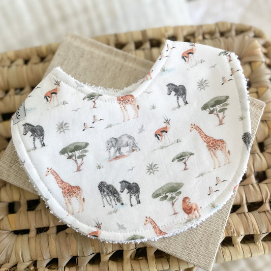 Flatlay of a handmade baby bib with a safari animal print including giraffes, elephants, zebras, birds, and trees on a soft neutral background. Made with cotton and white terry towelling backing.