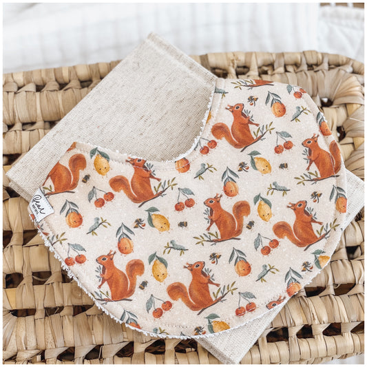 Close-up of a handmade baby bib with a whimsical woodland print, including red squirrels, lemons, cherries, and bees on a cream base. Bib is backed with beige terry towelling and placed on a woven basket.