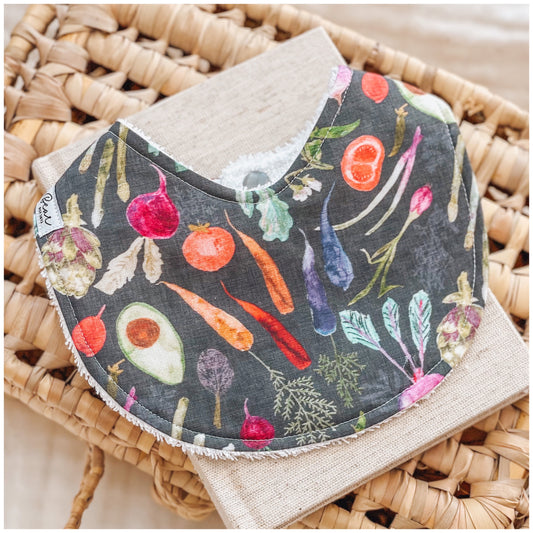 Baby bib with a charcoal background featuring assorted illustrated vegetables including carrots, radishes, avocado, onions, and leafy greens, styled on a rattan basket with neutral cloth underneath.