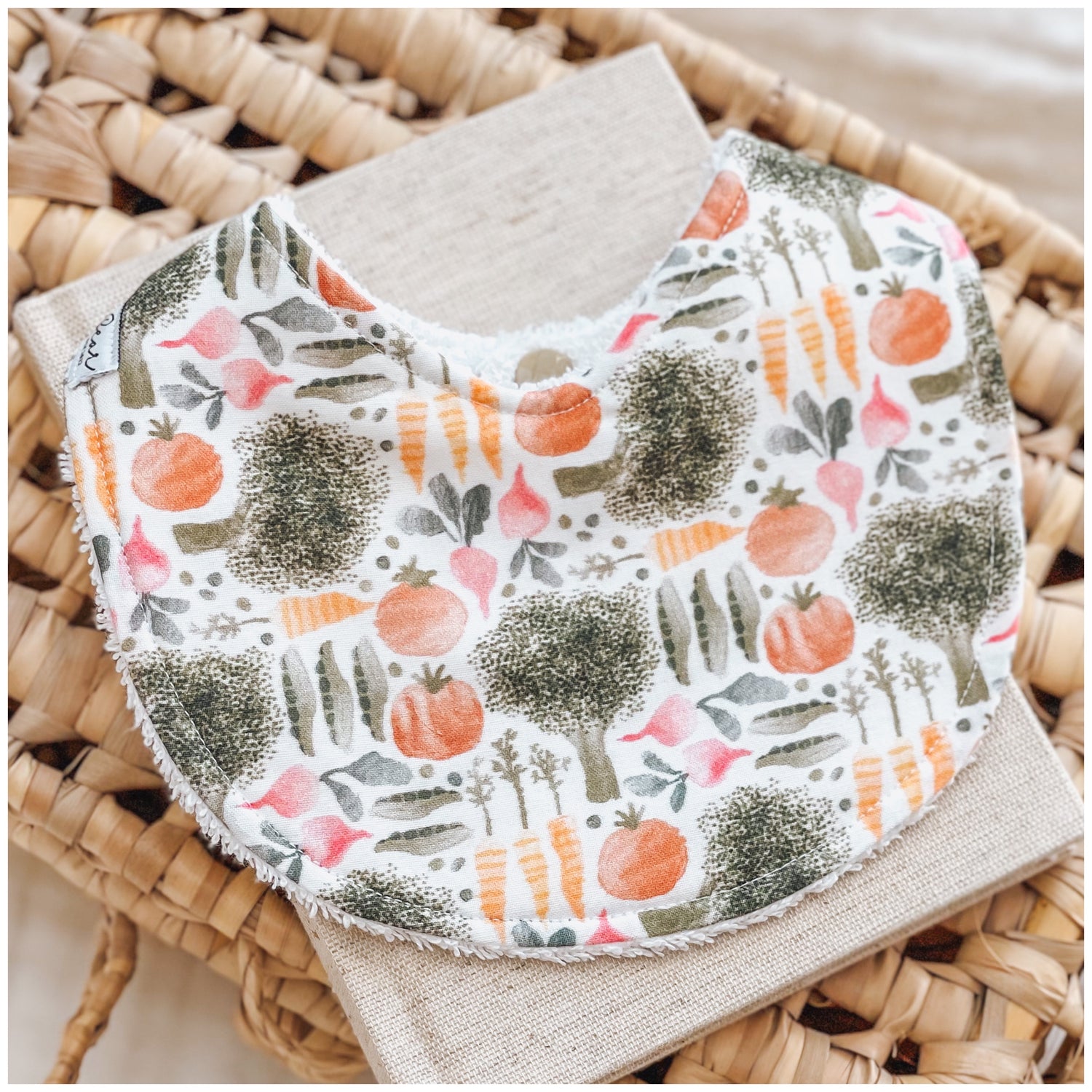 Baby bib with a garden vegetable print including broccoli, carrots, tomatoes, peas, and radishes on a white background. Bib is displayed on a woven basket with a neutral linen cloth beneath it.