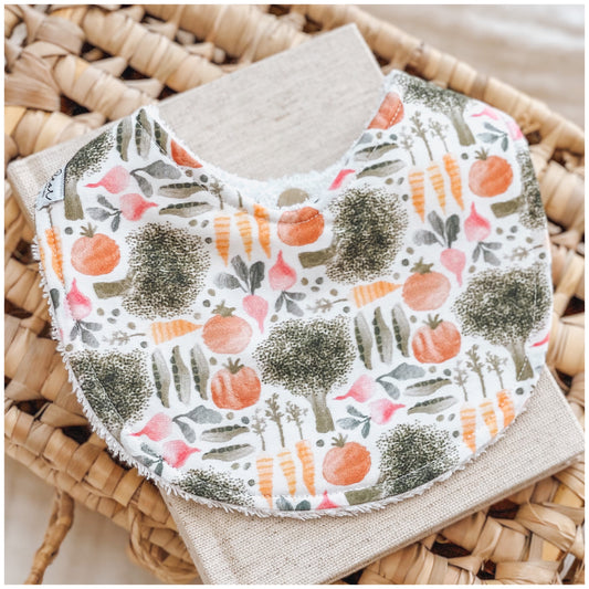 Baby bib with a garden vegetable print including broccoli, carrots, tomatoes, peas, and radishes on a white background. Bib is displayed on a woven basket with a neutral linen cloth beneath it.