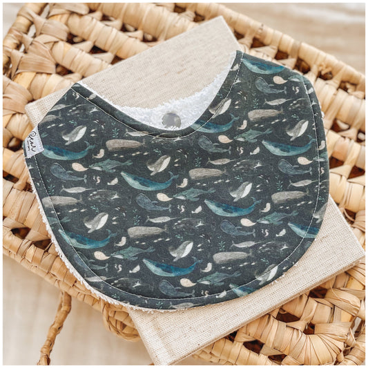 Handmade baby bib with a dark teal ocean background featuring illustrations of whales in soft blue, grey, and cream tones. Bib is styled on a woven basket with a natural linen cloth underneath.