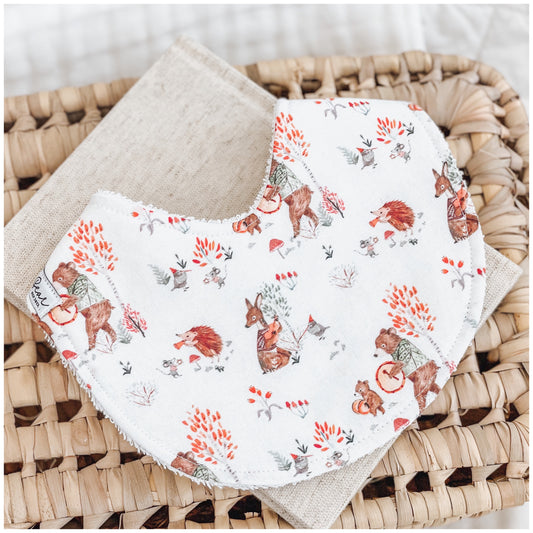 Neutral baby bib with playful woodland animals wearing scarves and jumpers among orange trees and mushrooms, displayed on a linen cloth and woven basket.