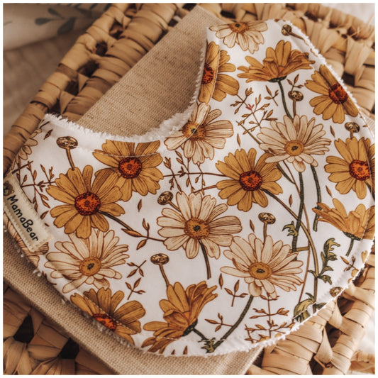 Handmade baby bib with a retro-inspired daisy print in mustard, beige, and rust on a white background, displayed on a piece of linen over a woven basket.