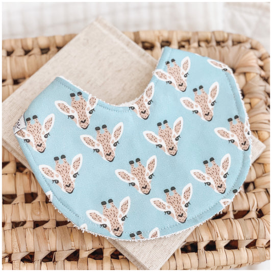 Handmade baby bib with a repeating giraffe face pattern on a bright sky blue background. Bib is backed with beige terry towelling and displayed on a woven basket.