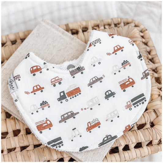 Handmade baby bib with a pattern of cars, trucks, and construction vehicles in rust, brown, and grey tones on a white background. Bib is backed with beige terry towelling and displayed on a woven basket.
