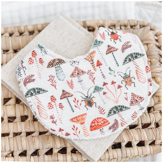 Handmade baby bib with a woodland pattern showing red, orange, and green mushrooms, forest leaves, and beetles on a soft neutral background. Bib is backed with beige terry towelling and styled on a woven basket.