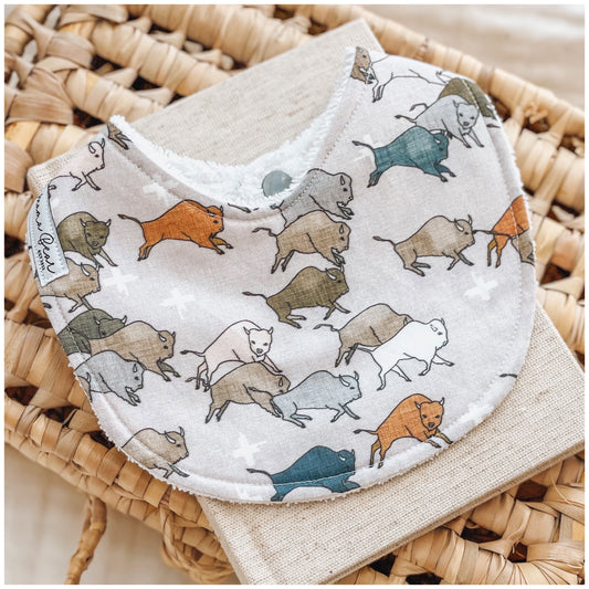 Baby bib with a playful print of illustrated buffalos in shades of brown, cream, teal, and orange on a soft beige background with white plus signs, displayed on a neutral linen cloth and woven tray.