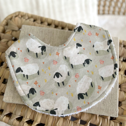 Baby bib with a soft green background featuring white sheep, orange and yellow wildflowers, and a terry towelling backing. Ideal for newborn feeding, dribble, and teething mess.