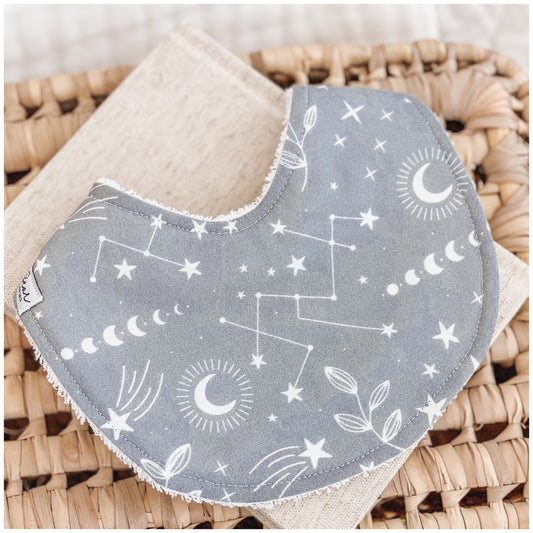 Handmade baby bib with a blue background and white celestial print, including stars, constellations, crescent moons, and leafy illustrations. Bib is backed with beige terry towelling and placed on a woven basket.
