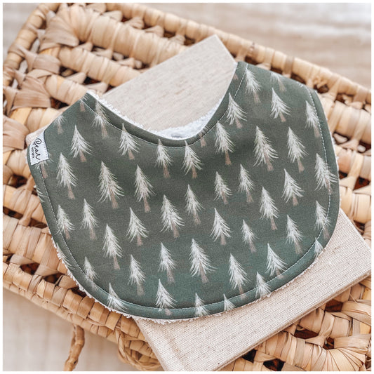 Baby bib with a forest green base and delicate white fir trees scattered across the fabric, styled on a woven basket with a neutral linen cloth