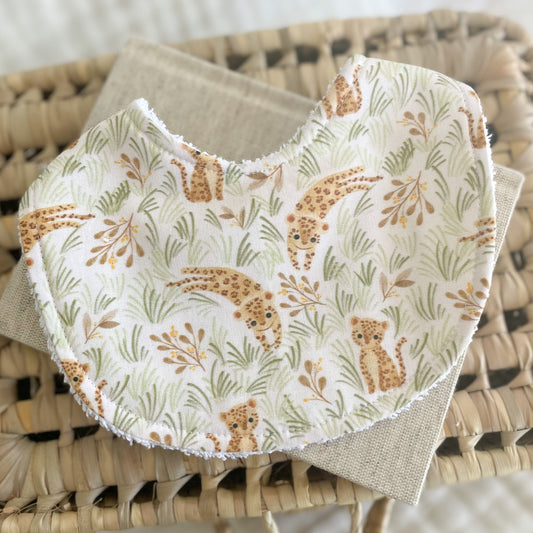 Baby bib with a cheetah cub pattern in soft earthy tones, surrounded by pale green grass and beige leaves. Displayed flat on a woven mat with a neutral fabric background.