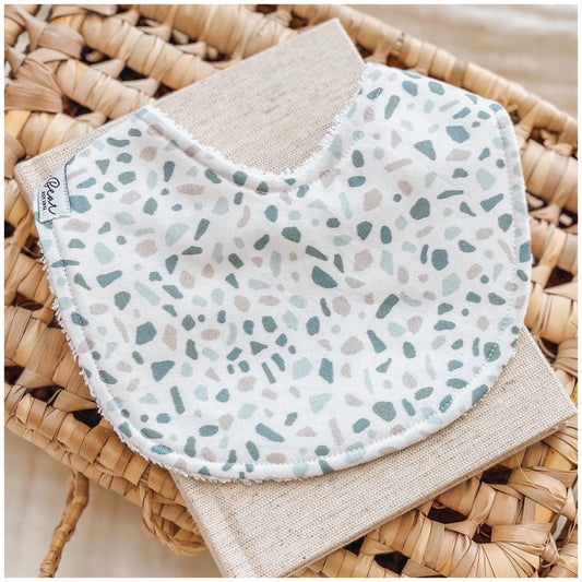 Baby bib featuring a terrazzo-style pattern with scattered sage green, blue, and beige speckles on a white background, displayed on a woven basket with a natural linen cloth underneath.