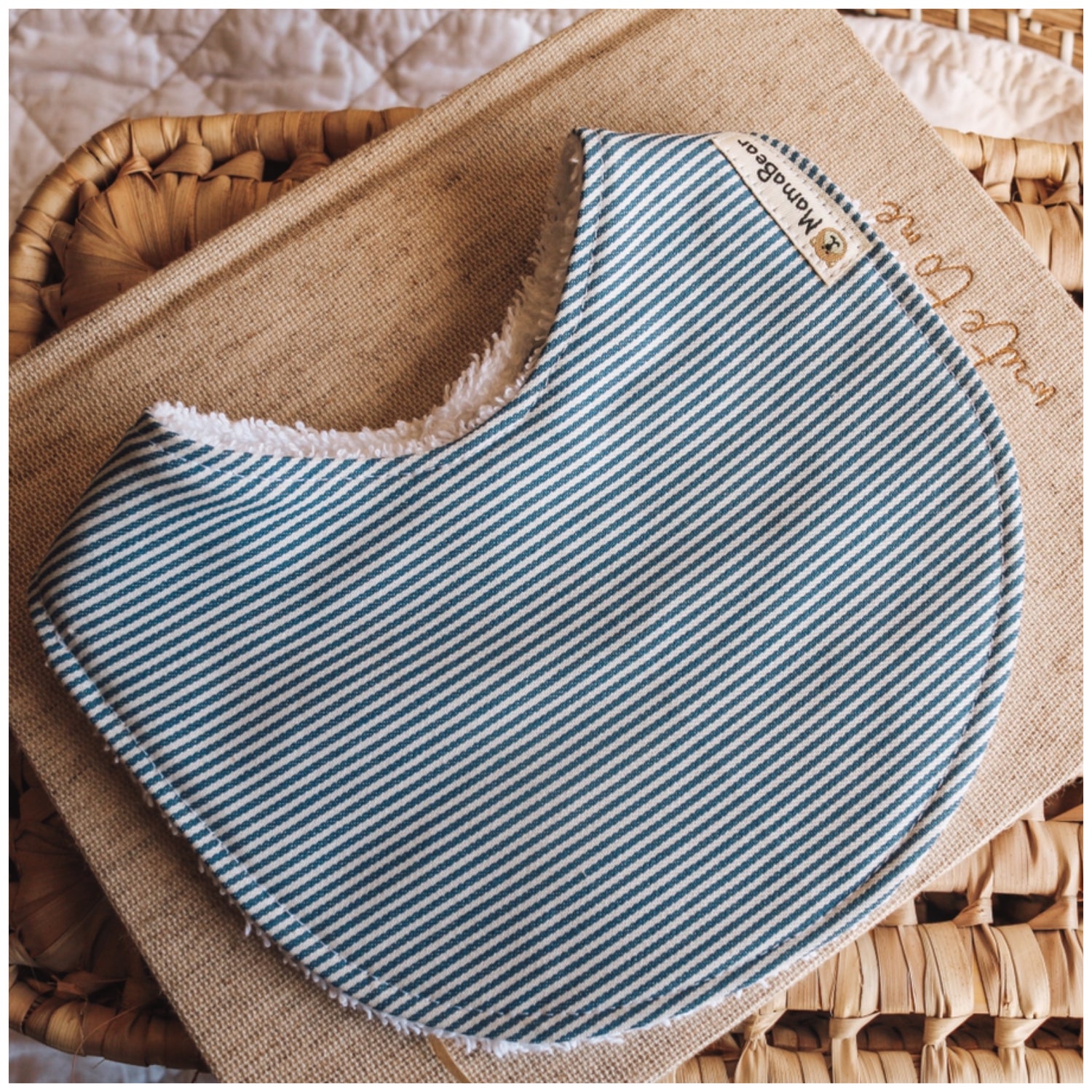 Handmade baby bib with a classic blue and white pinstripe pattern, backed with white terry towelling, styled on natural fabric and a woven basket background.