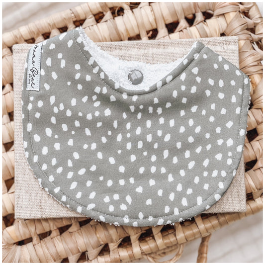 Sage green baby bib with an all-over scattered white spot pattern, styled on a neutral background with a woven basket.