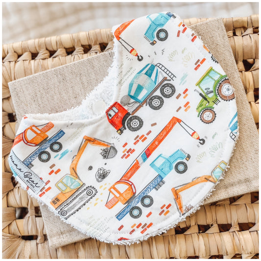 Baby bib with a colourful construction theme, featuring illustrated diggers, dump trucks, cement mixers, and cranes on a white background. Displayed on a woven basket with a neutral cloth — a fun and practical baby gift for truck-loving families.