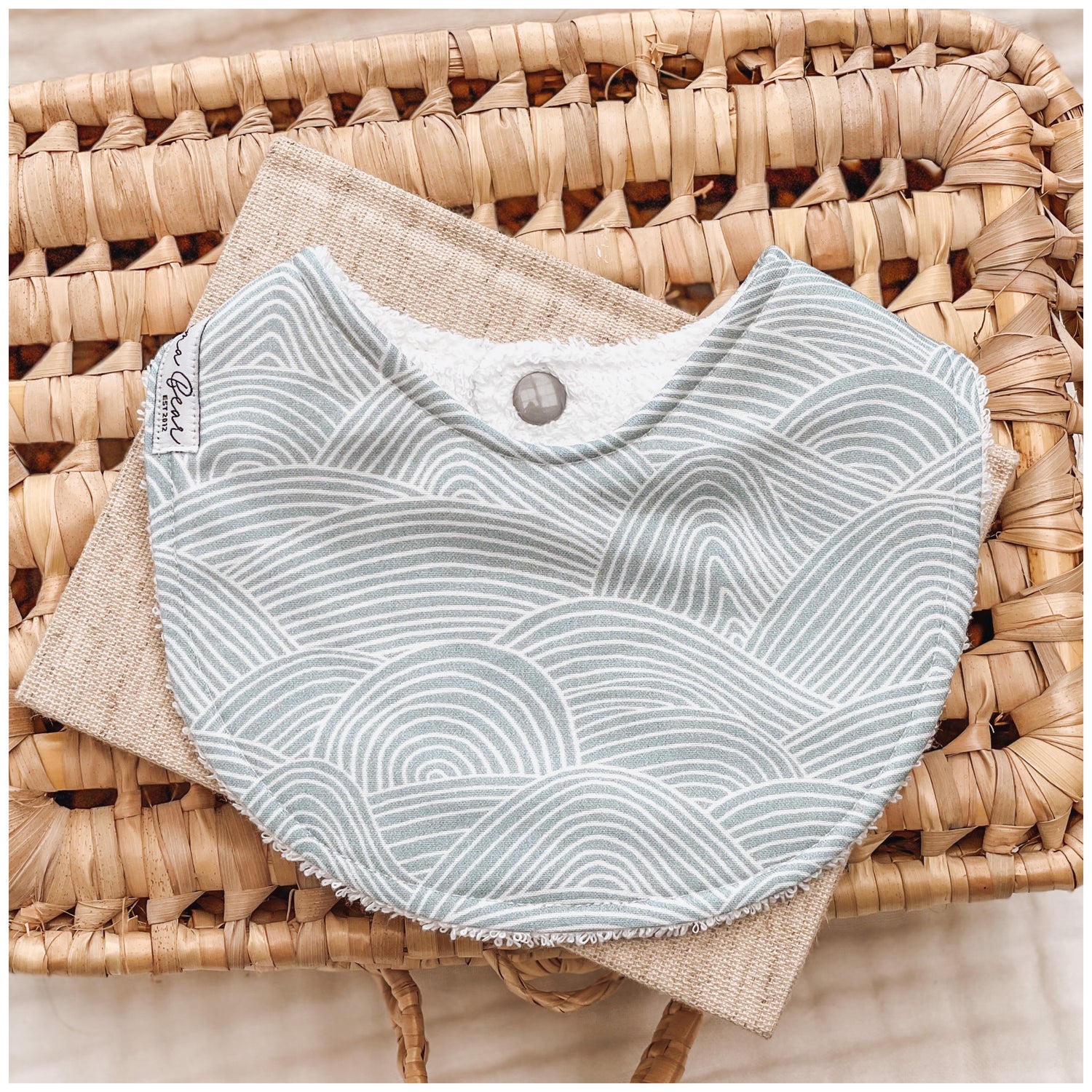 Seafoam blue baby bib featuring white curved line waves in a layered, ripple-style pattern, resting on neutral-toned fabric in a wicker basket.