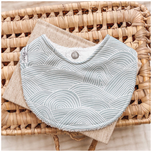 Seafoam blue baby bib featuring white curved line waves in a layered, ripple-style pattern, resting on neutral-toned fabric in a wicker basket.