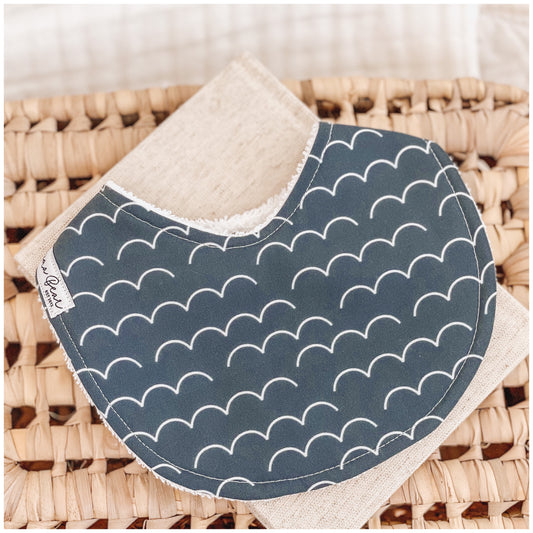 Handmade baby bib featuring a deep navy blue background with a repeating white scalloped arch pattern, placed on a woven surface.