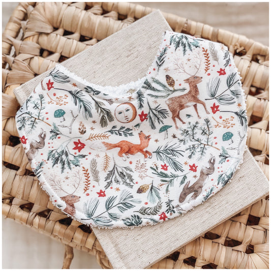 Woodland-themed baby bib with illustrated deer, foxes, squirrels, blue shaded pine needles, red leaves, and scattered forest elements on a soft white background.