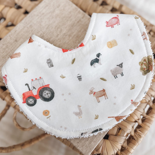 Farm-themed baby bib with scattered illustrations of red tractors, pigs, sheep, dogs, goats, hay bales, and chickens on a white background. Lined with terry towelling for absorbency.