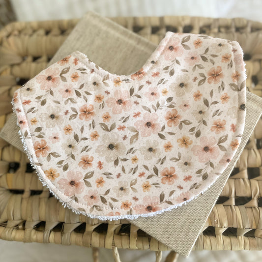 Floral baby bib with blush, apricot, and beige flowers on a cream background, displayed on a neutral woven basket. Ideal for newborns, dribble, and teething.