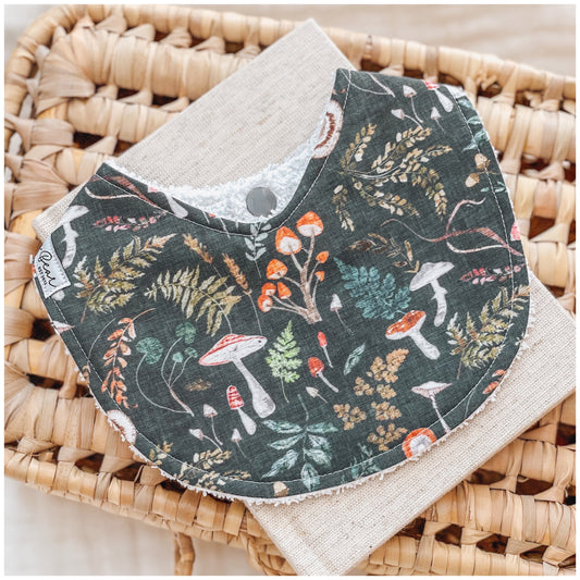Dark green baby bib with a whimsical forest pattern of orange and red mushrooms, leafy ferns, and woodland plants. A gender-neutral design perfect for nature-loving babies.
