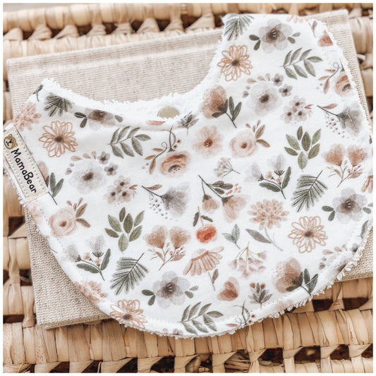 Baby bib with a woodland floral print in warm earthy tones, including soft browns, greens, and muted pinks. Photographed on a woven basket with a natural linen cloth — a neutral, handmade baby bib ideal for newborns.