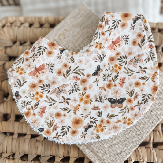 Handmade baby bib featuring a delicate nature-inspired print with butterflies, bees, leafy branches, and soft wildflowers in peach, mustard, and earthy brown tones. Set on a white background and backed with absorbent terry towelling, perfect for dribbles and teething.