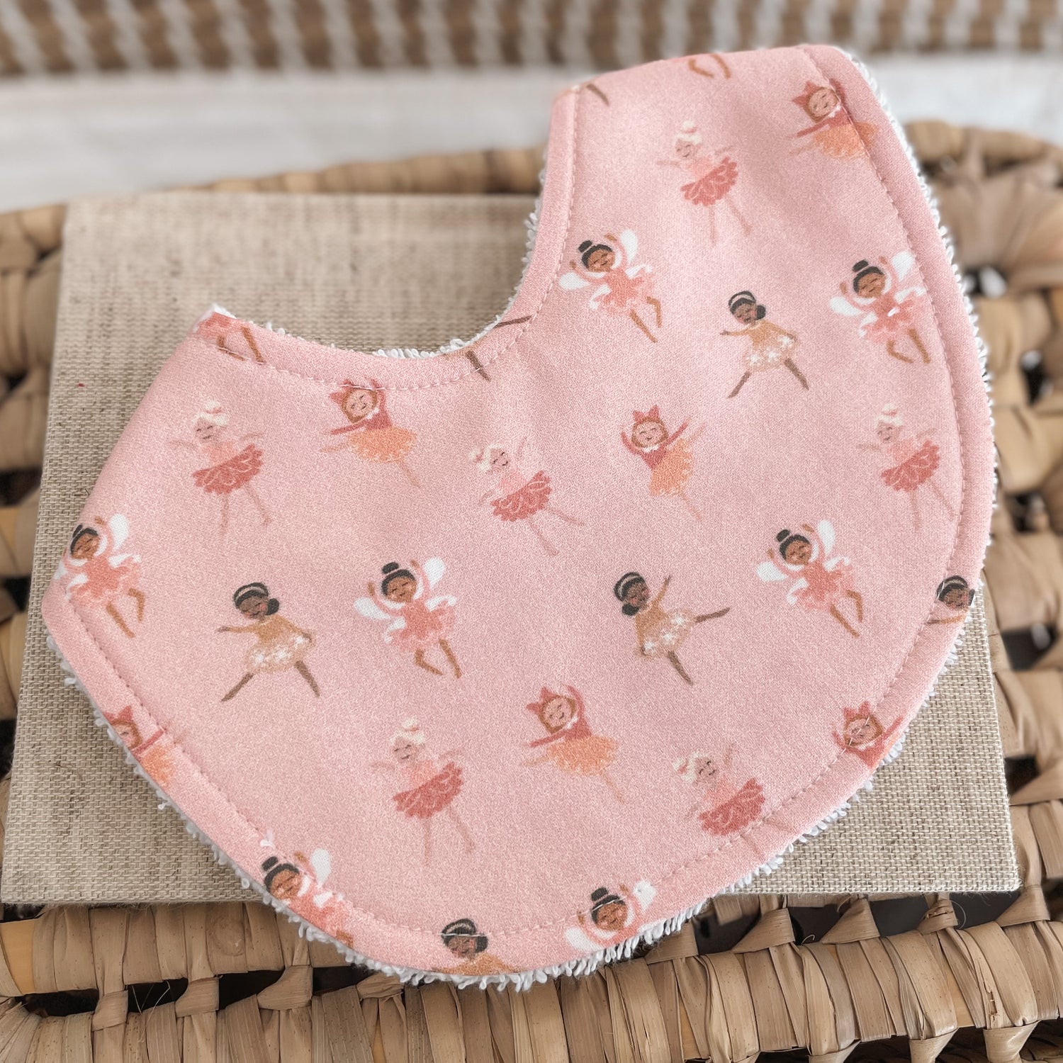 Flatlay of a handmade baby bib with a pink fairy ballerina print. The design shows tiny fairies and ballerinas in various dance poses wearing tutus and wings, scattered across a soft blush-pink background. The bib is backed with absorbent white terry towelling.