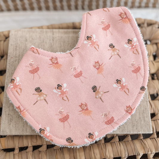 Flatlay of a handmade baby bib with a pink fairy ballerina print. The design shows tiny fairies and ballerinas in various dance poses wearing tutus and wings, scattered across a soft blush-pink background. The bib is backed with absorbent white terry towelling.