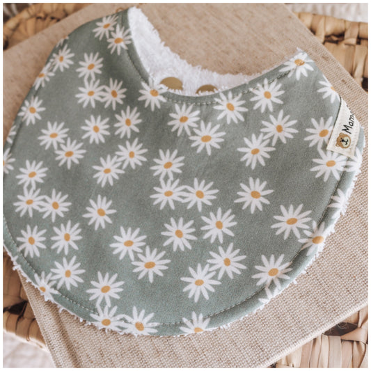 Handmade baby bib with white daisy flowers and mustard centres on a sage green background, backed with soft white terry towelling and styled on natural fabric.
