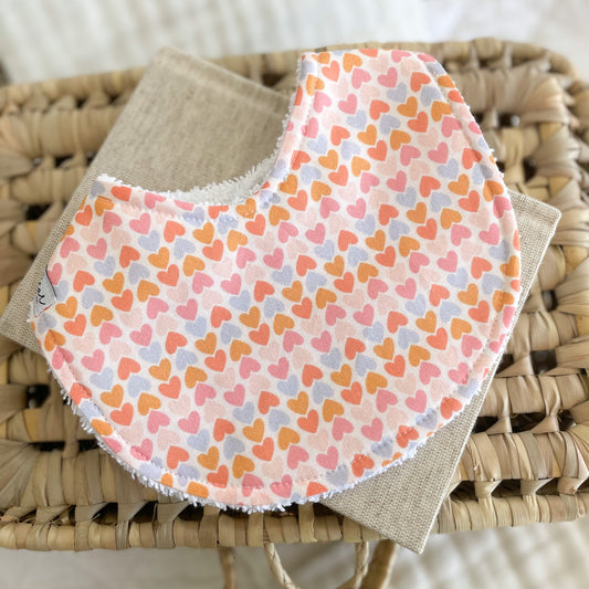 Flatlay of a handmade baby bib featuring a repeating pattern of small hearts in rainbow shades of pink, peach, orange and lilac on a soft white background. Bib has a cotton front and white terry towelling back for absorbency.