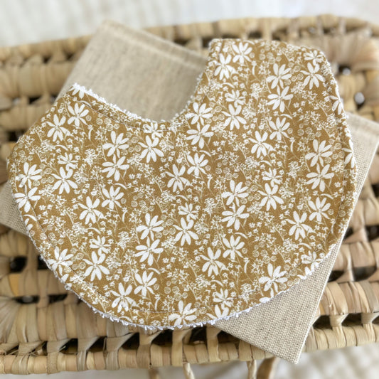 Flatlay of a handmade baby bib with a mustard yellow background and small white floral print. The flowers are scattered closely across the fabric, giving it a vintage look. Bib is made from cotton with white terry towelling backing.