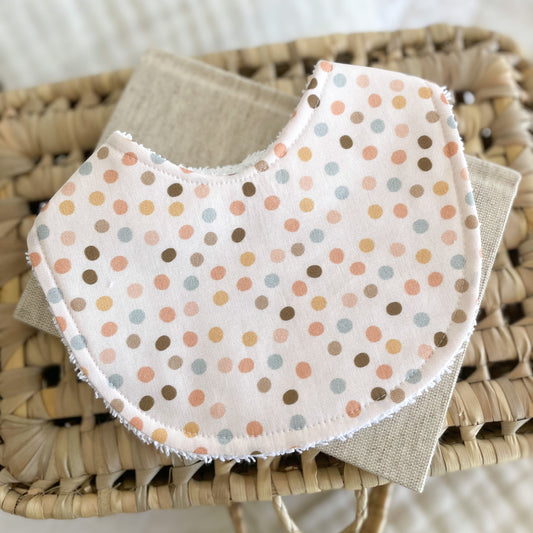 Handmade baby bib with pastel pink, blue, and mustard polka dots on a cream background. Made with cotton front and terry towelling backing, shown flatlay on woven surface.
