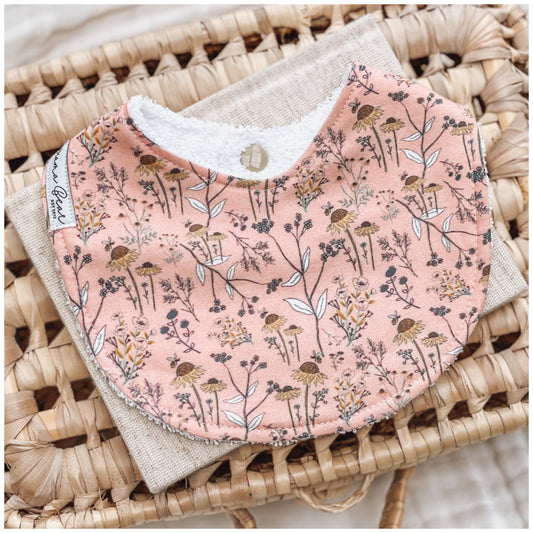 Peach-coloured baby bib with an all-over print of delicate wildflowers in mustard, white, and dusty mauve, handmade with absorbent terry towelling backing.