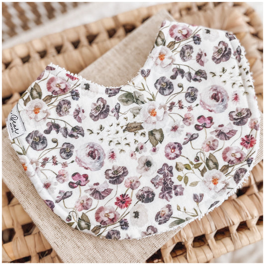 Baby bib with a soft white background featuring an all-over print of delicate purple, mauve, and lavender watercolour-style flowers, backed with white terry towelling.