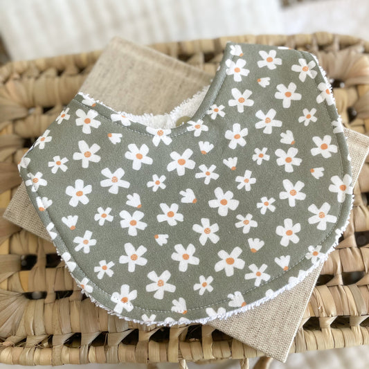 Flatlay of a handmade baby bib with a sage green background and white daisy flower print. The flowers have orange centres, giving the design a soft, natural feel. The bib is made from cotton with a terry towelling reverse side for absorbency.