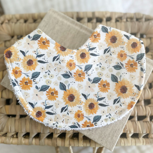 Handmade baby bib featuring a sunflower print with golden yellow and soft beige flowers on a white background, styled on neutral-toned fabric and a woven basket.