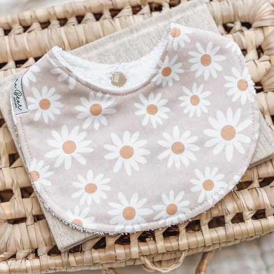 Handmade baby bib with a playful white daisy floral print on a beige background, displayed on a woven mat with neutral-toned props.