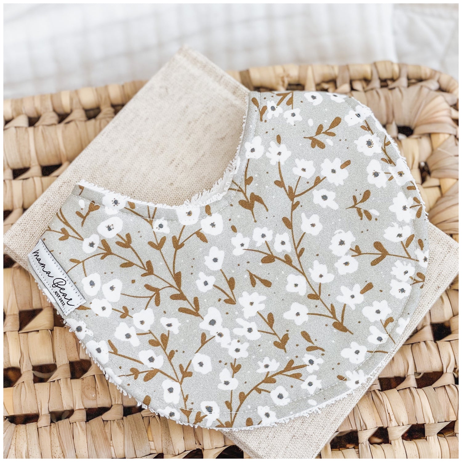 Baby bib featuring white dainty floral sprigs with golden brown stems on a muted sage grey background, displayed on a linen cloth and wicker basket.