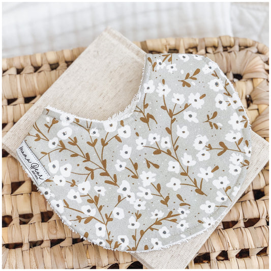 Baby bib featuring white dainty floral sprigs with golden brown stems on a muted sage grey background, displayed on a linen cloth and wicker basket.
