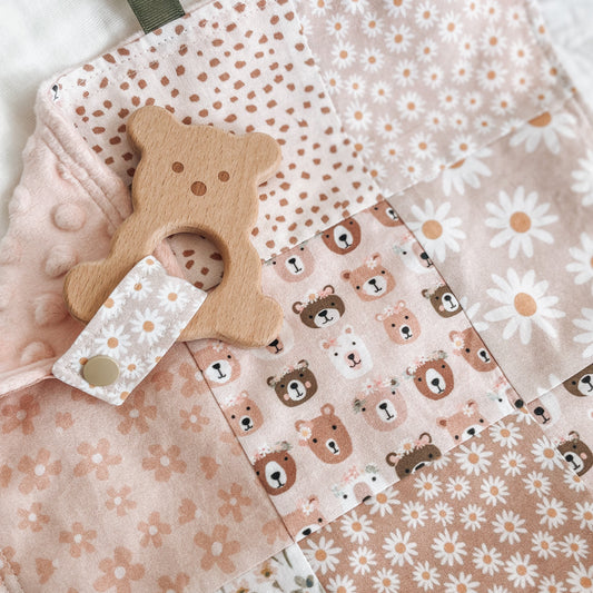 Close-up of a handmade baby Taggie blankie in soft pink and neutral tones, featuring patchwork squares with daisies, teddy bears, florals, and polka dots. Finished with textured minkee backing, ribbon loops, and a wooden bear-shaped teether attached with a floral fabric strap.