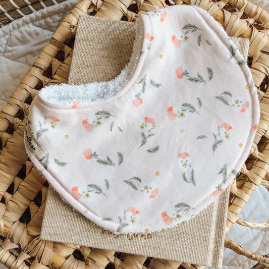 Handmade baby bib in soft blush pink cotton with tiny floral clusters in peach, white, and sage green, backed with absorbent white towelling and styled on natural fabric and woven basket.