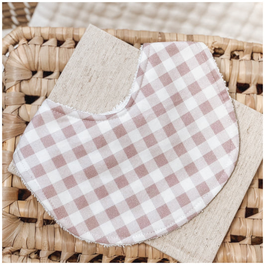 Baby bib in a soft lilac and white gingham check pattern, photographed on a woven basket with a neutral fabric backing. Handmade and perfect for newborns, this bib is a sweet and practical baby gift idea.
