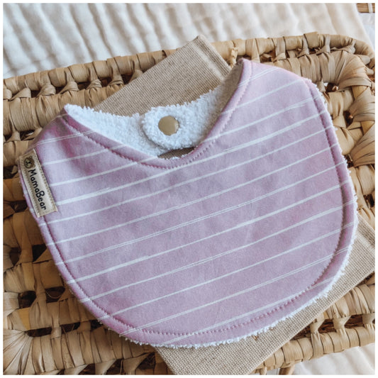 Baby bib in soft lilac with fine white horizontal stripes. Displayed on a woven basket with a natural fabric base — a handmade baby girl bib in a simple and timeless design.