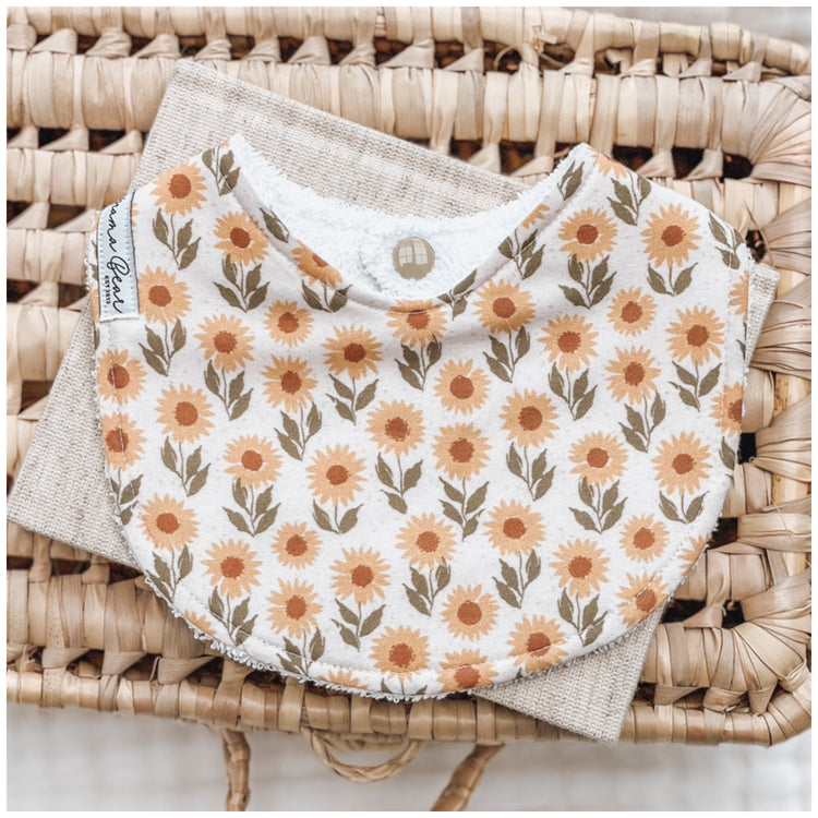 Cream baby bib with scattered orange sunflower print, displayed on a woven basket with a neutral linen cloth underneath.