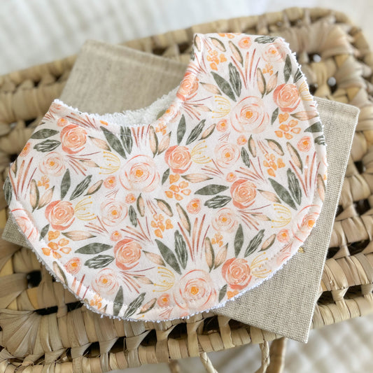 Flatlay of a handmade baby bib with a romantic floral pattern. The design includes pink and peach flowers with soft green leaves on a light cream base. Bib is made from cotton with an absorbent white terry towelling back.