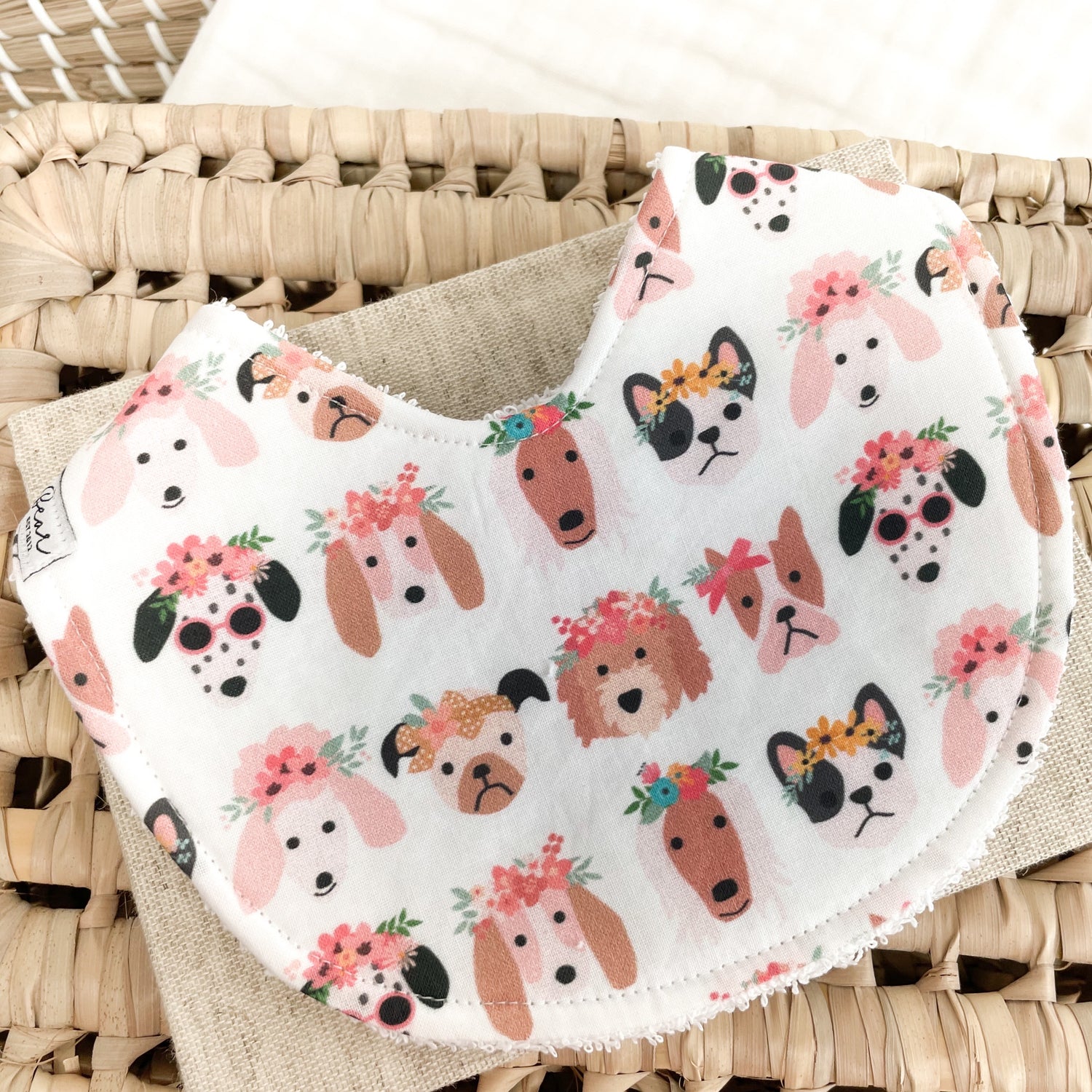 White baby bib with a fun dog print—featuring cartoon-style puppies wearing flower crowns, bows, and sunglasses. Absorbent terry towelling backing, ideal for teething and feeding.